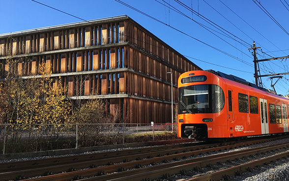 Ein oranger RBS-Zug fährt am BAV-Gebäude vorbei, das eine Holzfassade hat.
